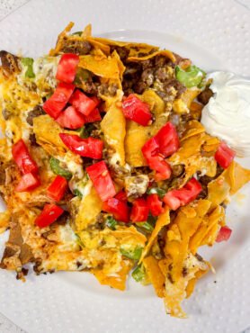 Keto nachos piled with cheese, celery, tomatoes and a side of sour cream on a white plate.
