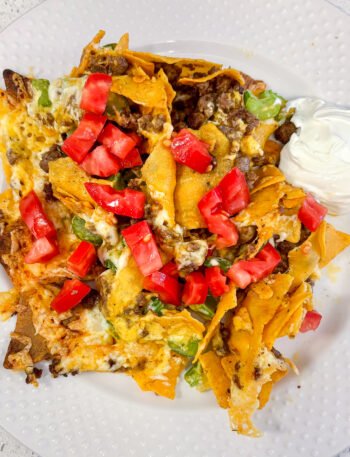Keto nachos piled with cheese, celery, tomatoes and a side of sour cream on a white plate.