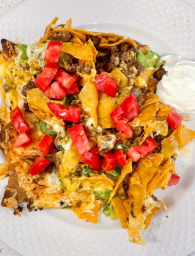 Keto nachos piled with cheese, celery, tomatoes and a side of sour cream on a white plate.